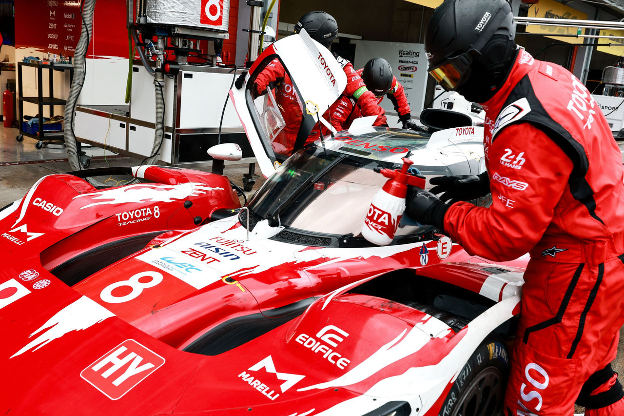 Pit lane scene showing a red-and-white Toyota endurance race car, number 8, stopped for service while multiple crew members in protective gear work on the cockpit area.